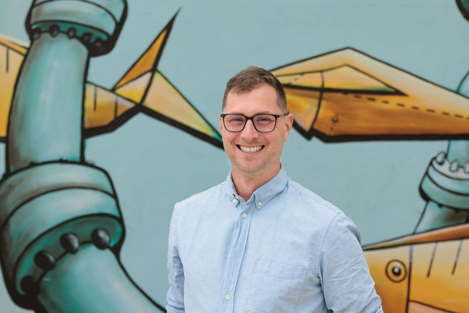 A profile photo featuring Tim McNamara prominently. Tim is standing in front of a painted wall featuring an artwork that has urban themes, including pipes and freshwater.